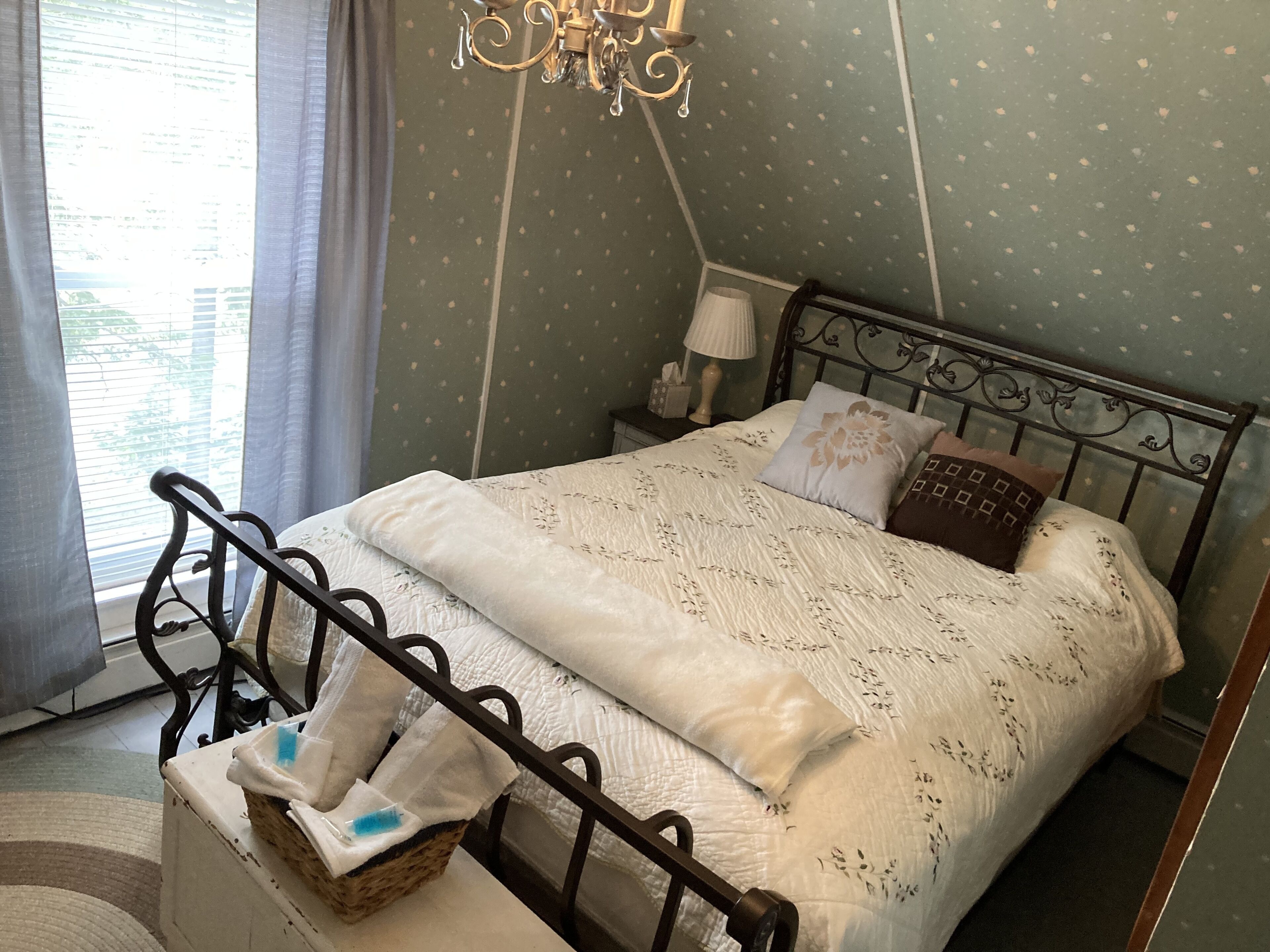 Un lit bien fait, recouvert d’une couette blanche et agrémenté de coussins décoratifs, une table de chevet avec une lampe, et une fenêtre avec des rideaux.