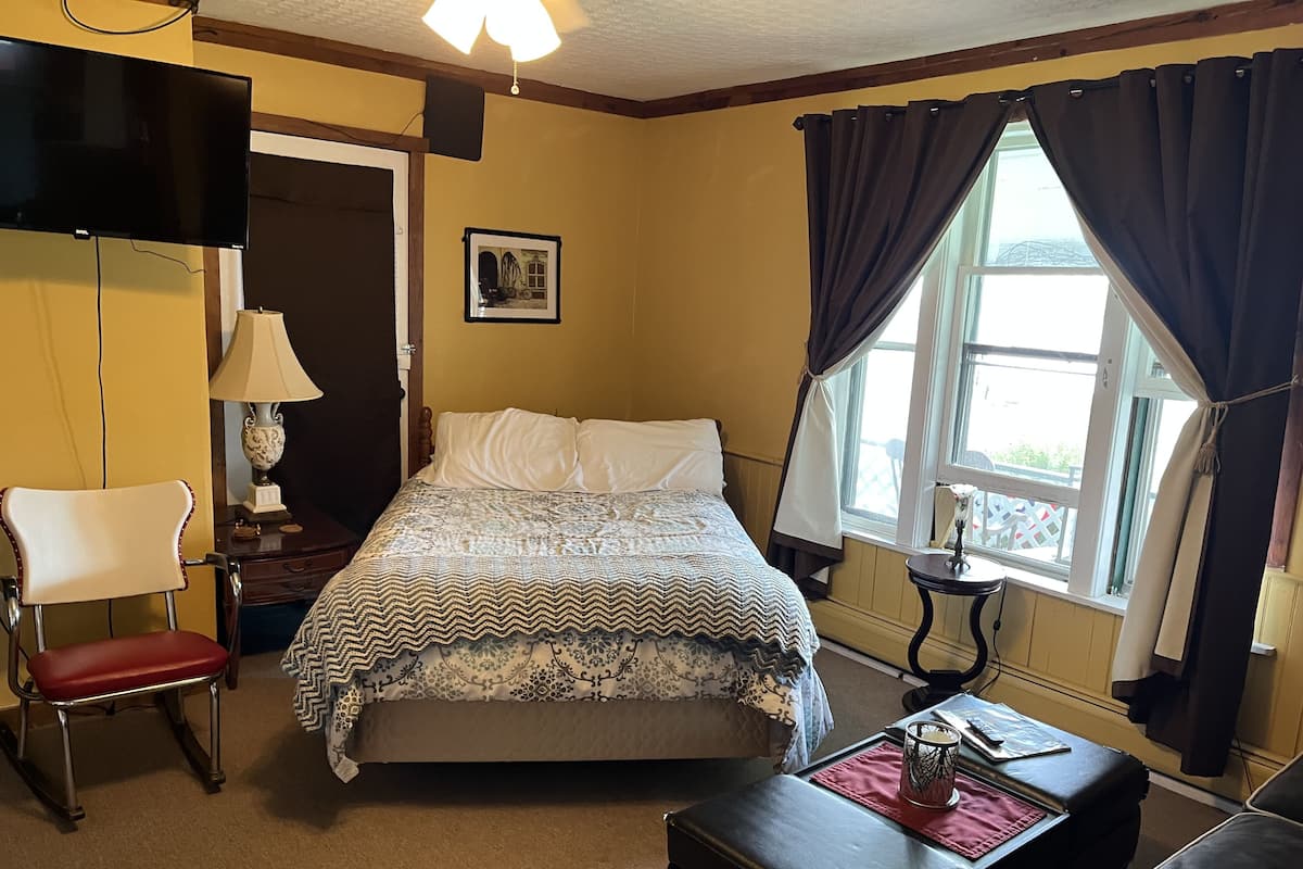 Un dormitorio con cama, una silla, escritorio, lámpara, un cuadro en la pared y una ventana con cortinas.
