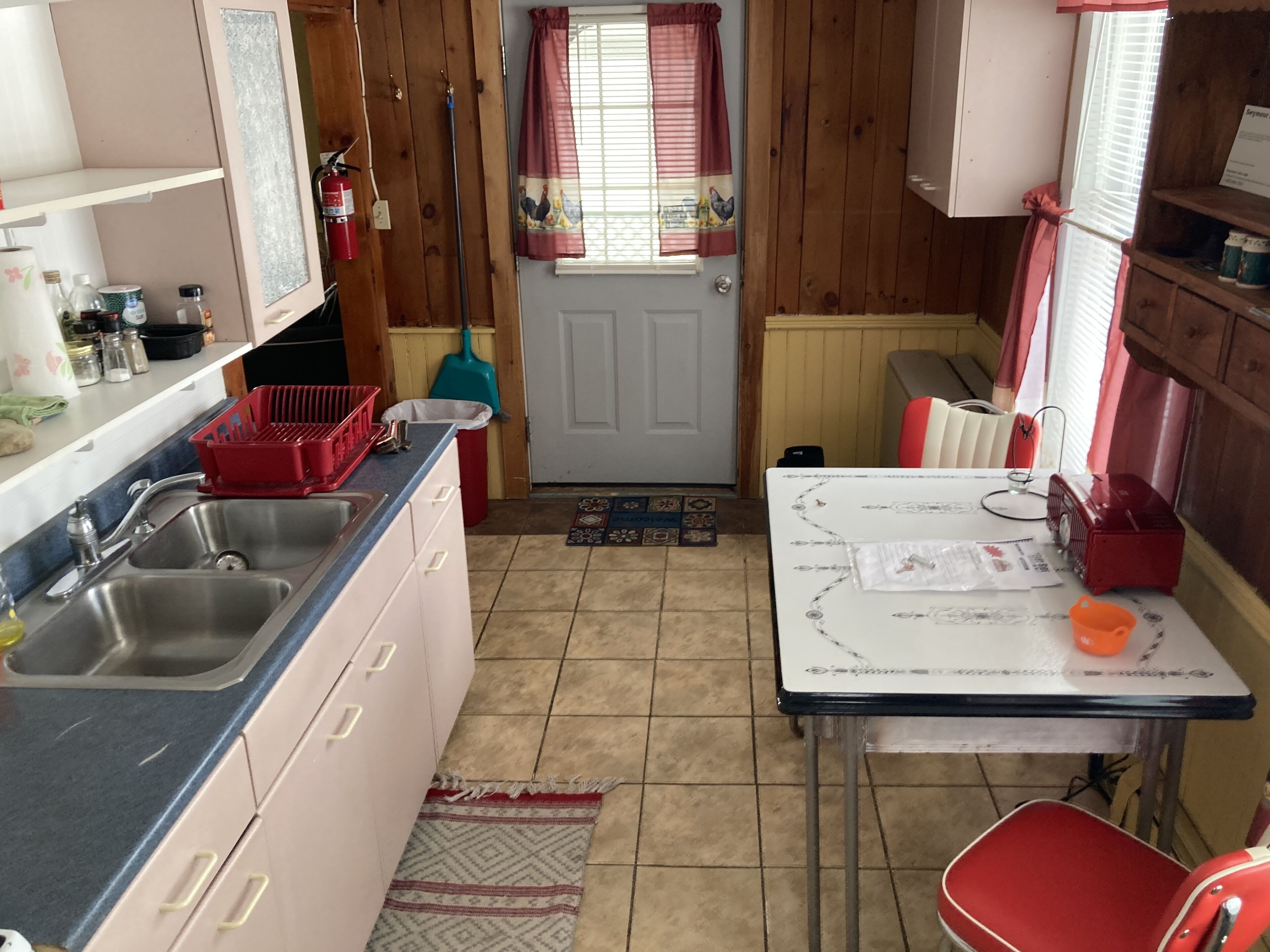 A compact kitchen with a sink, a table, and a chair. There is a window with curtains and a door.
