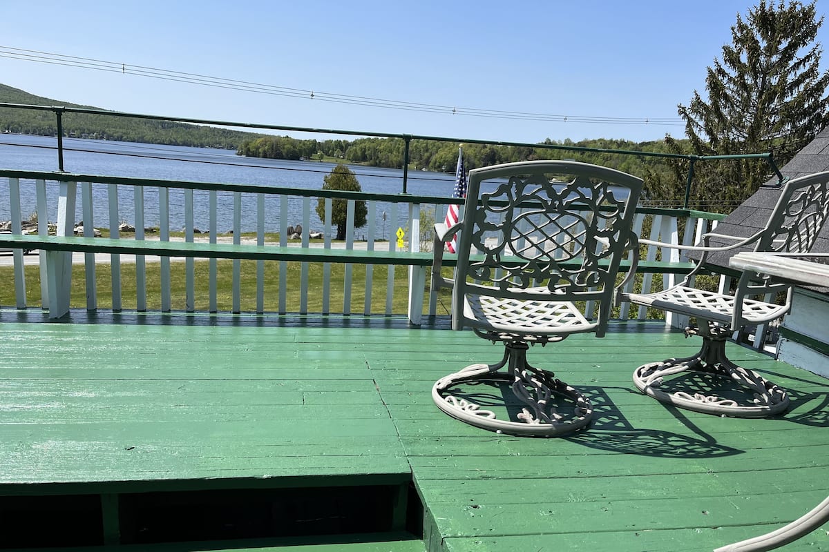 Una terraza verde con dos sillas ornamentales y una mesa, con vistas a un lago que incluye una cerca blanca y árboles al fondo.