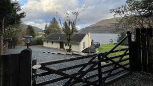 Une maison avec une allée en gravier et un portail en bois, offrant une vue sur un lac et des collines.