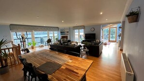 Un vaste espace de vie comprenant une table à manger en bois, des canapés et un téléviseur. De grandes fenêtres offrent une vue sur l’extérieur.