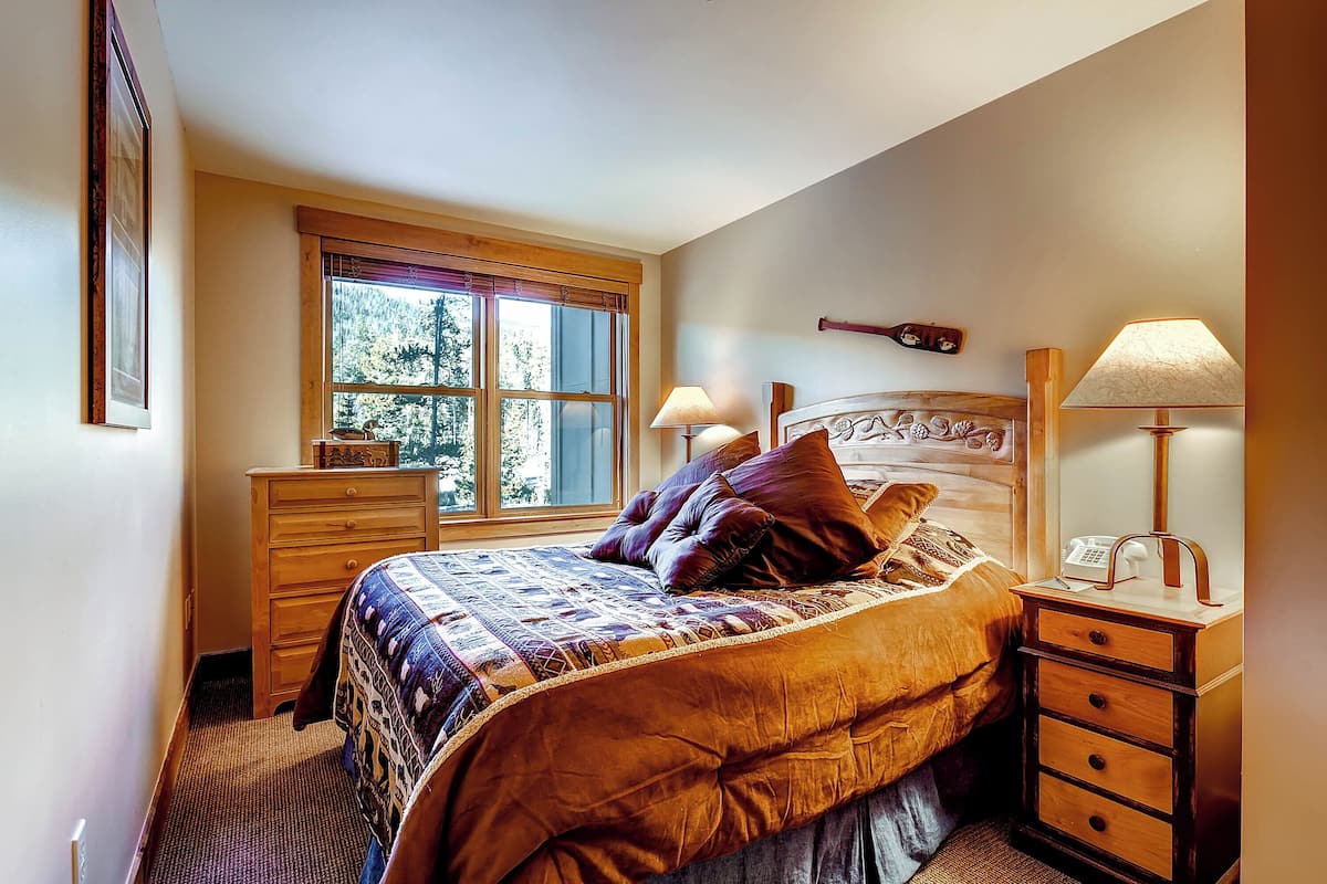 A bedroom with a large bed, a dresser, a nightstand with lamps, and a window with a view of trees.