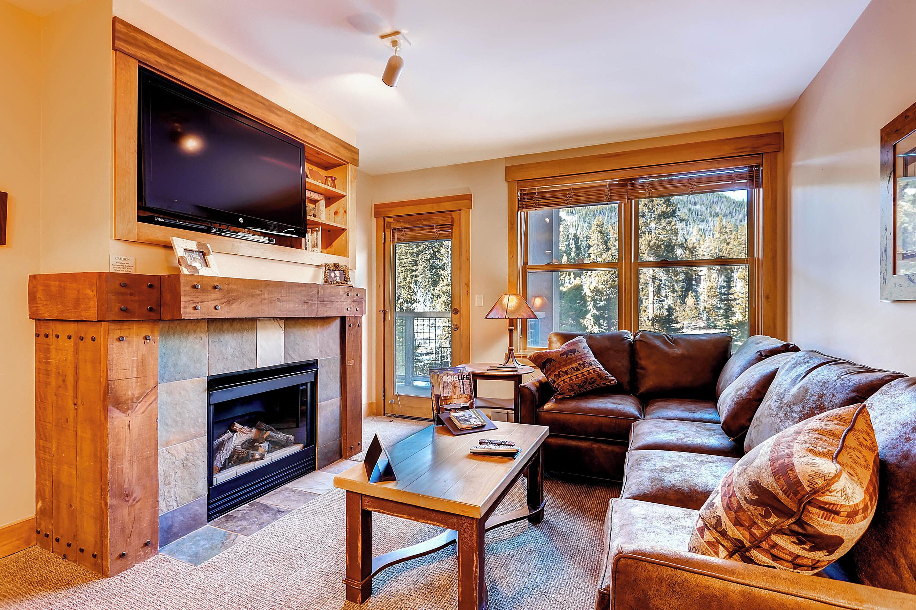 A cozy living room with a fireplace, a large flat-screen TV mounted above it, a leather sofa, and a coffee table.