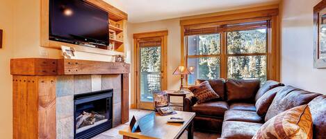 A cozy living room with a fireplace, a large flat-screen TV mounted above it, a leather sofa, and a coffee table.