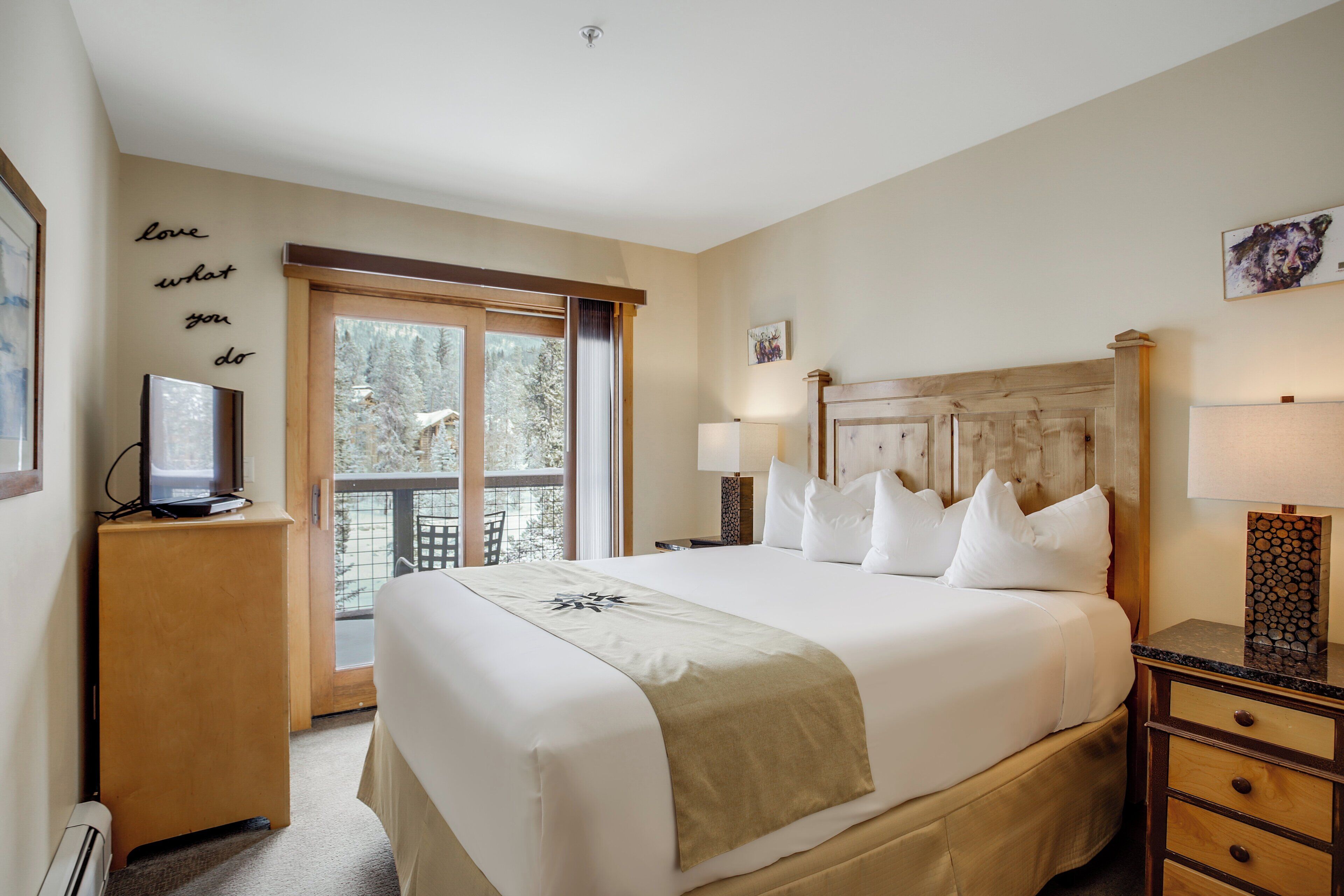 Une chambre d’hôtel avec un grand lit, un téléviseur posé sur un meuble, un balcon donnant sur la verdure et un mur orné d’une citation inspirante.