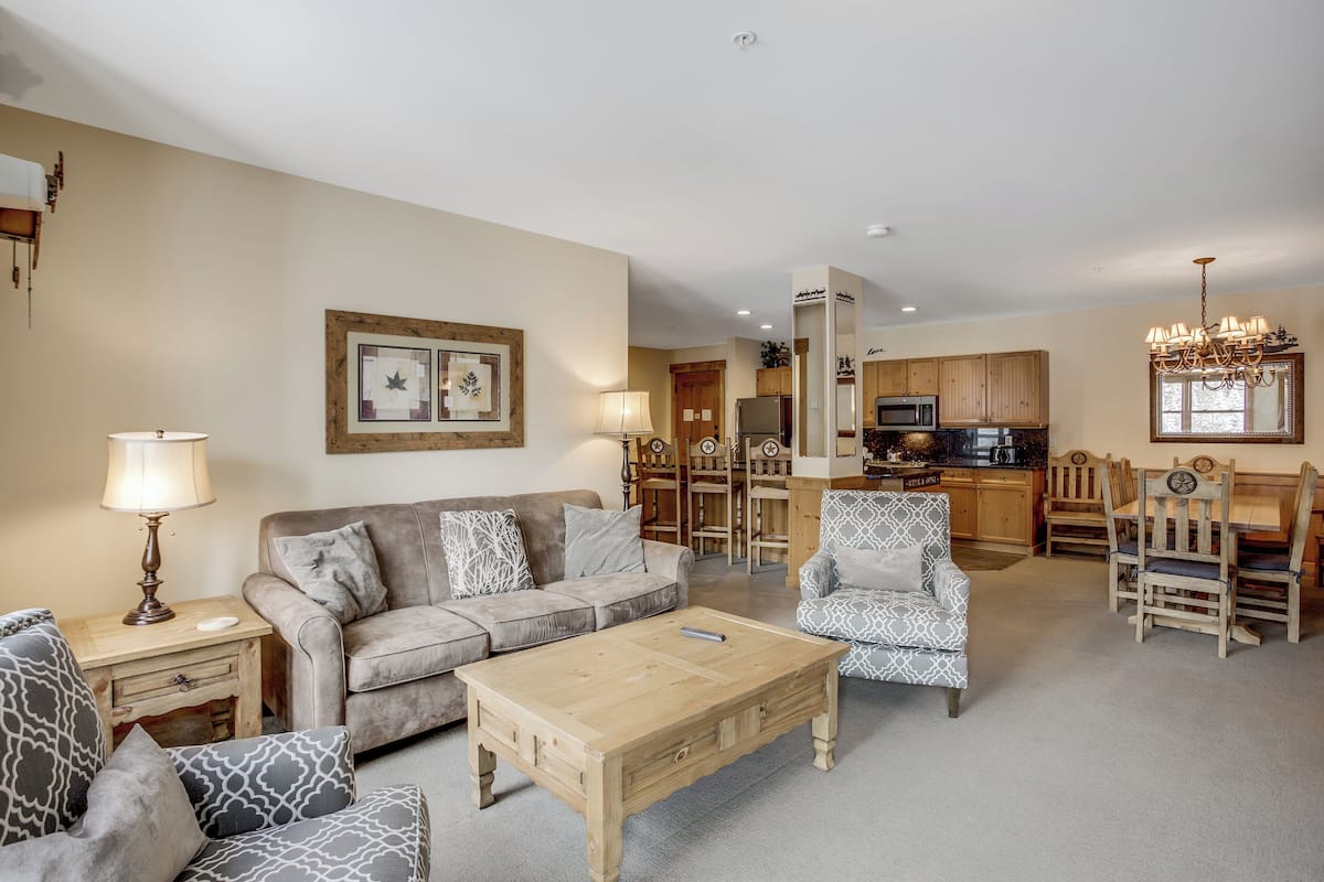 A spacious living room with a sofa, coffee table, patterned chairs, and a dining area with wooden chairs and a chandelier.