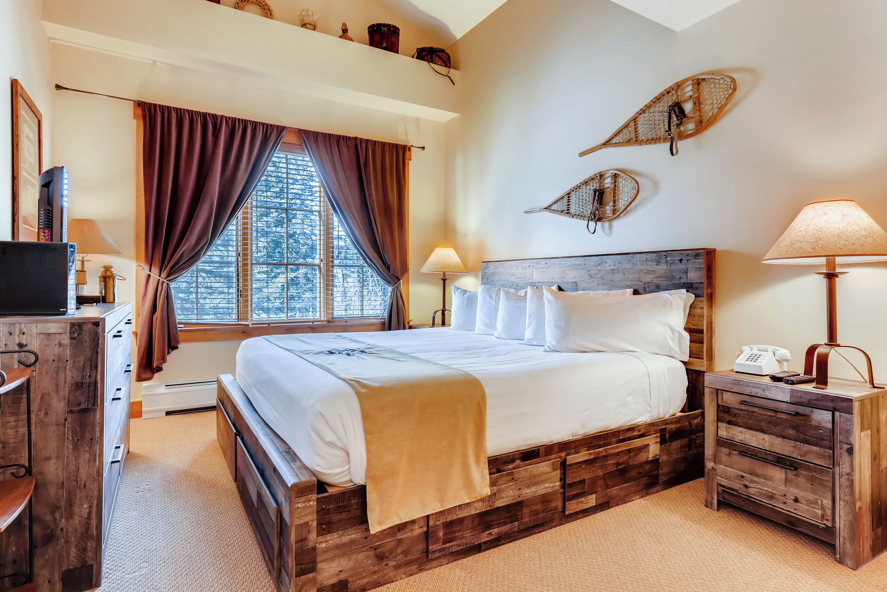A bedroom with a wooden bed, a desk, a chair, a window with curtains, and a wall-mounted shelf.