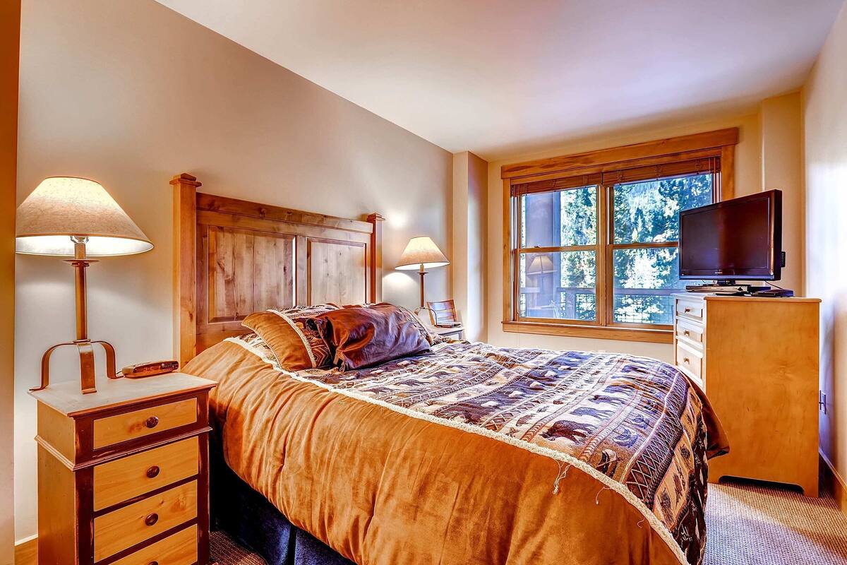 A bedroom with a large bed, wooden headboard, bedside lamps, a dresser, a television, and a window with a view of trees.