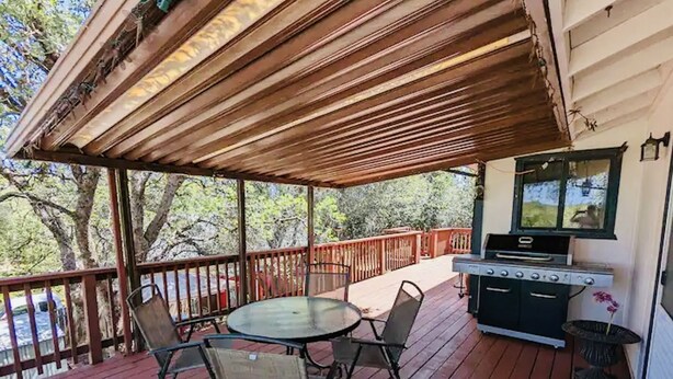 A covered outdoor deck with a grill, a table with chairs, and a view of trees.