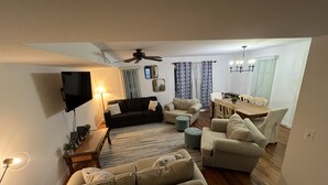 A living room with a sofa, ottoman, and a TV. There is a dining area with a table and chairs. - Family Friendly Townhome in desired Stewards Watch Community- 5mins to the beach (Bethany Beach)