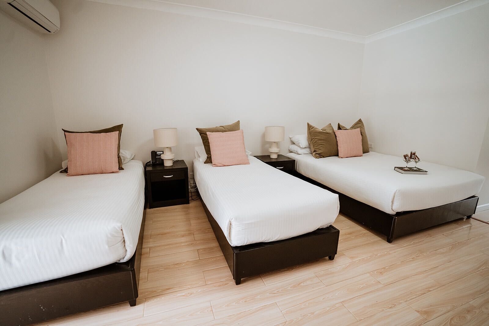 A hotel room with three single beds, each with a bedside table and lamp. The room has wooden flooring and white walls.