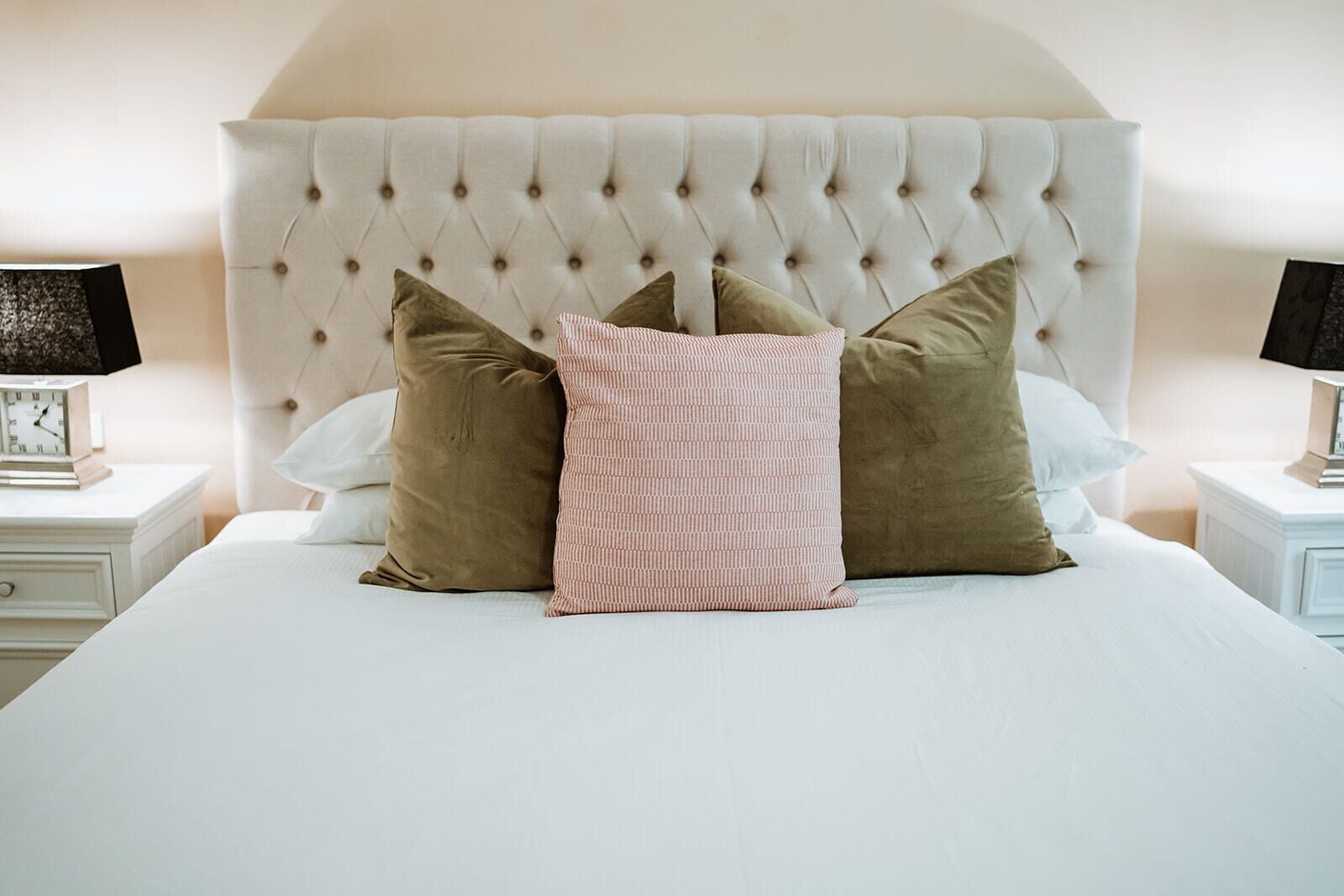 A neatly made bed with a tufted headboard, two bedside tables with lamps, and a clock on one of the tables.