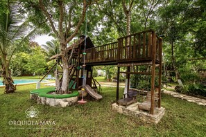 Une structure de jeux en bois, dotée d’un toboggan et d’une zone d’escalade, entourée d’une végétation luxuriante et d’une piscine.