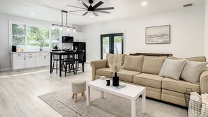 Ein modernes Wohnzimmer mit einem beigefarbenen Sofa, einem weißen Couchtisch und einem Essbereich mit einem schwarzen Tisch und Stühlen.