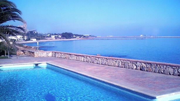 Une piscine à l’eau d’un bleu cristallin, un mur de pierre et une vue sur la mer et les bâtiments éloignés.