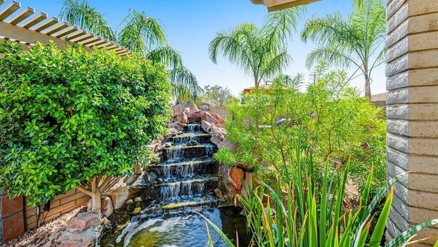 A waterfall in a landscaped garden with a stone wall and a pergola structure. - Backyard Oasis Ideal for Spring/Summer! Pool, Misters, Bluetooth Speakers + more (Mesa)