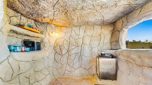 A stone-walled room with shelves holding various items, a small refrigerator, and a welcome mat. - Backyard Oasis Ideal for Spring/Summer! Pool, Misters, Bluetooth Speakers + more (Mesa)