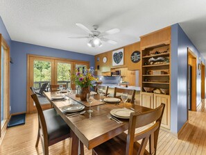 A dining area with a wooden table set for four, a ceiling fan, and a kitchen area with wooden cabinets and a clock.