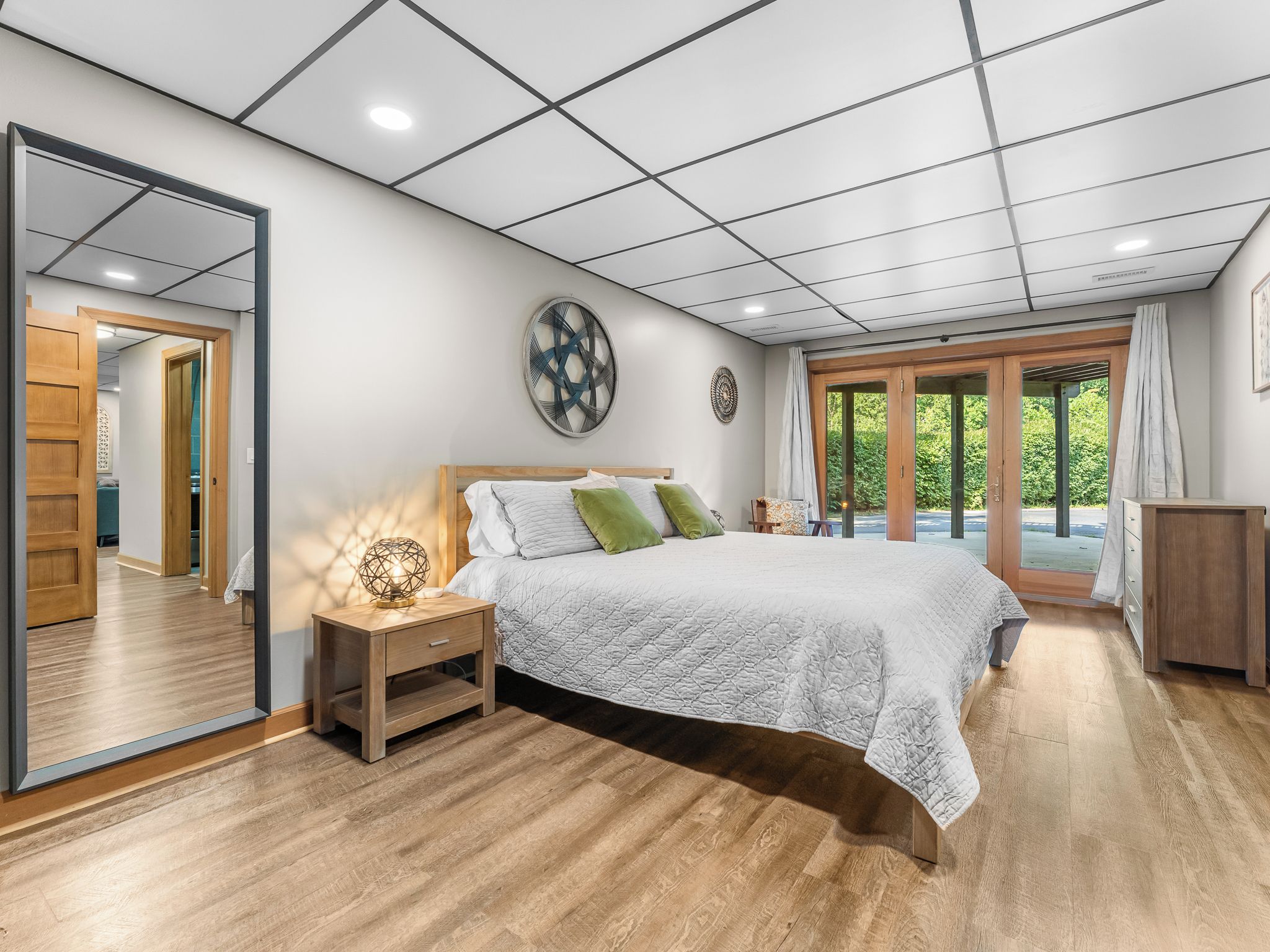 A bedroom with a large bed, a nightstand, a mirror, and a view of the outdoors through a glass door.