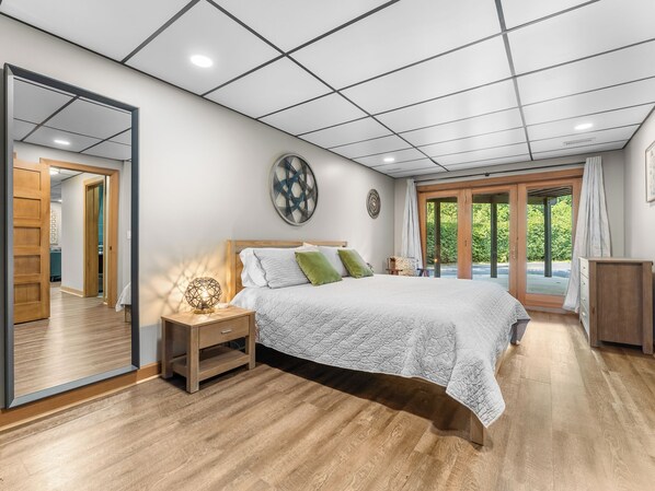 A bedroom with a large bed, a nightstand, a mirror, and a view of the outdoors through a glass door.