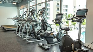 Un gimnasio con cintas de correr, elípticas y una bicicleta estática, con vista a un edificio y una piscina al aire libre.