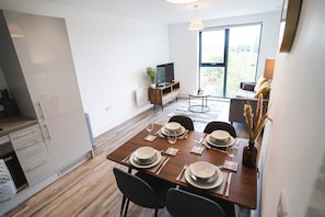 Uma área de refeições moderna com uma mesa de madeira posta para quatro pessoas, uma televisão em um móvel e uma janela grande com vista para o exterior.