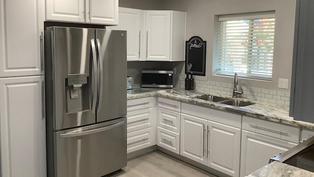 A modern kitchen with white cabinets, stainless steel appliances, a granite countertop, and a window with blinds. - Flawless Faith (Redding)