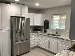 A modern kitchen with white cabinets, stainless steel appliances, a granite countertop, and a window with blinds.
