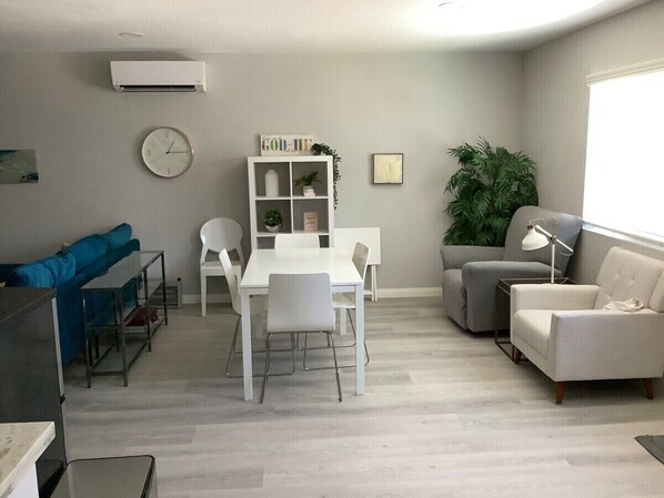 A modern living room with a white dining table, white chairs, a blue sofa, a grey sofa, and a grey armchair. There is a white shelf with plants and a clock on the wall.