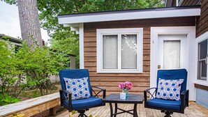 Eine Holzterrasse mit zwei blauen Stühlen und einem Tisch, einer kleinen Vase mit Blumen sowie einem Haus mit zwei Fenstern und einer Tür.