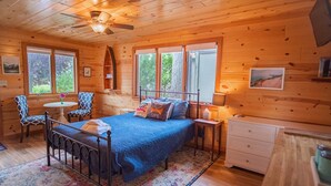 Ein Schlafzimmer in einer Holzhütte mit einem Bett, zwei Stühlen, einem kleinen Tisch, einer Kommode und einem Deckenventilator.