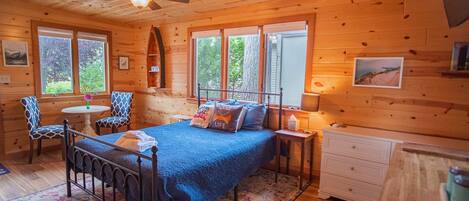 Un dormitorio de cabaña de madera con cama, dos sillas, una mesita, un aparador y un ventilador de techo.