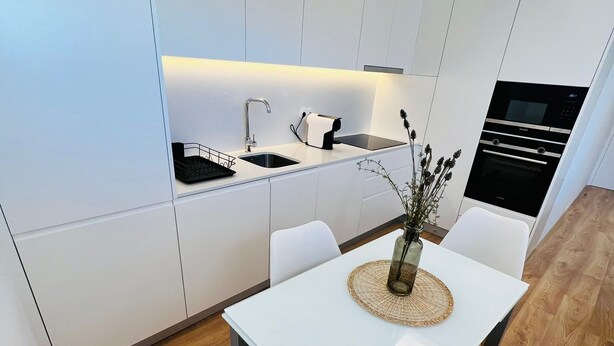 A modern kitchen with white cabinetry, a built-in oven, and a dining area with a white table and chairs.
