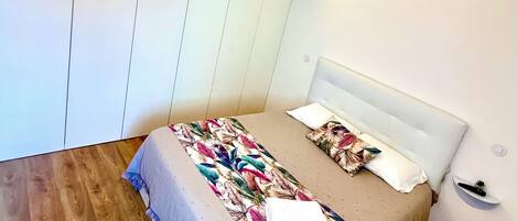 A bedroom with a bed, a nightstand, a wall-mounted shelf, and a wooden floor.