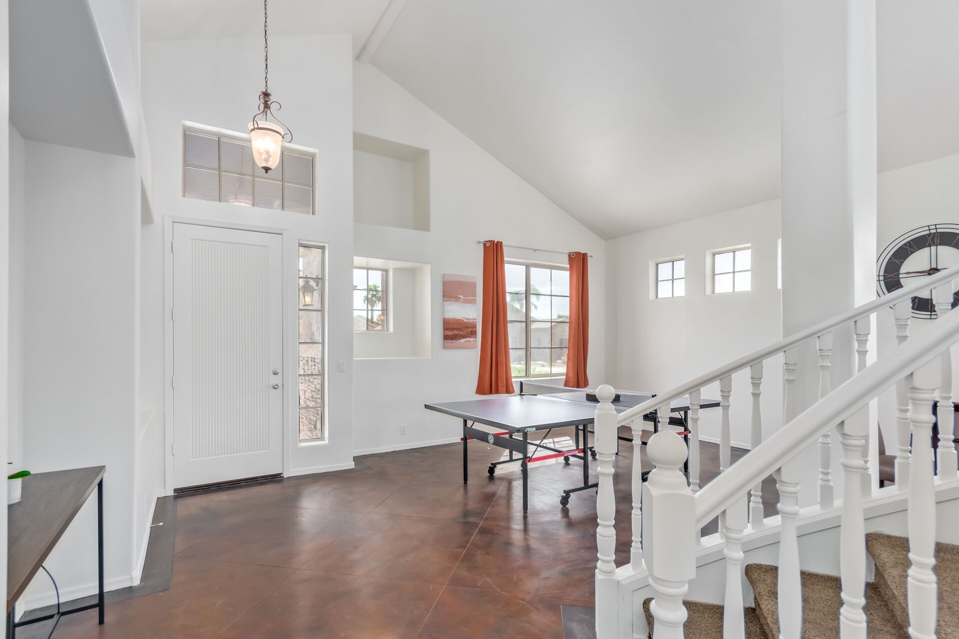 A spacious room with a ping pong table, a white staircase, and a large window with orange curtains.