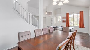 Un comedor amplio con una gran mesa de madera y seis sillas tapizadas, una escalera con barandillas blancas y un moderno candelabro.