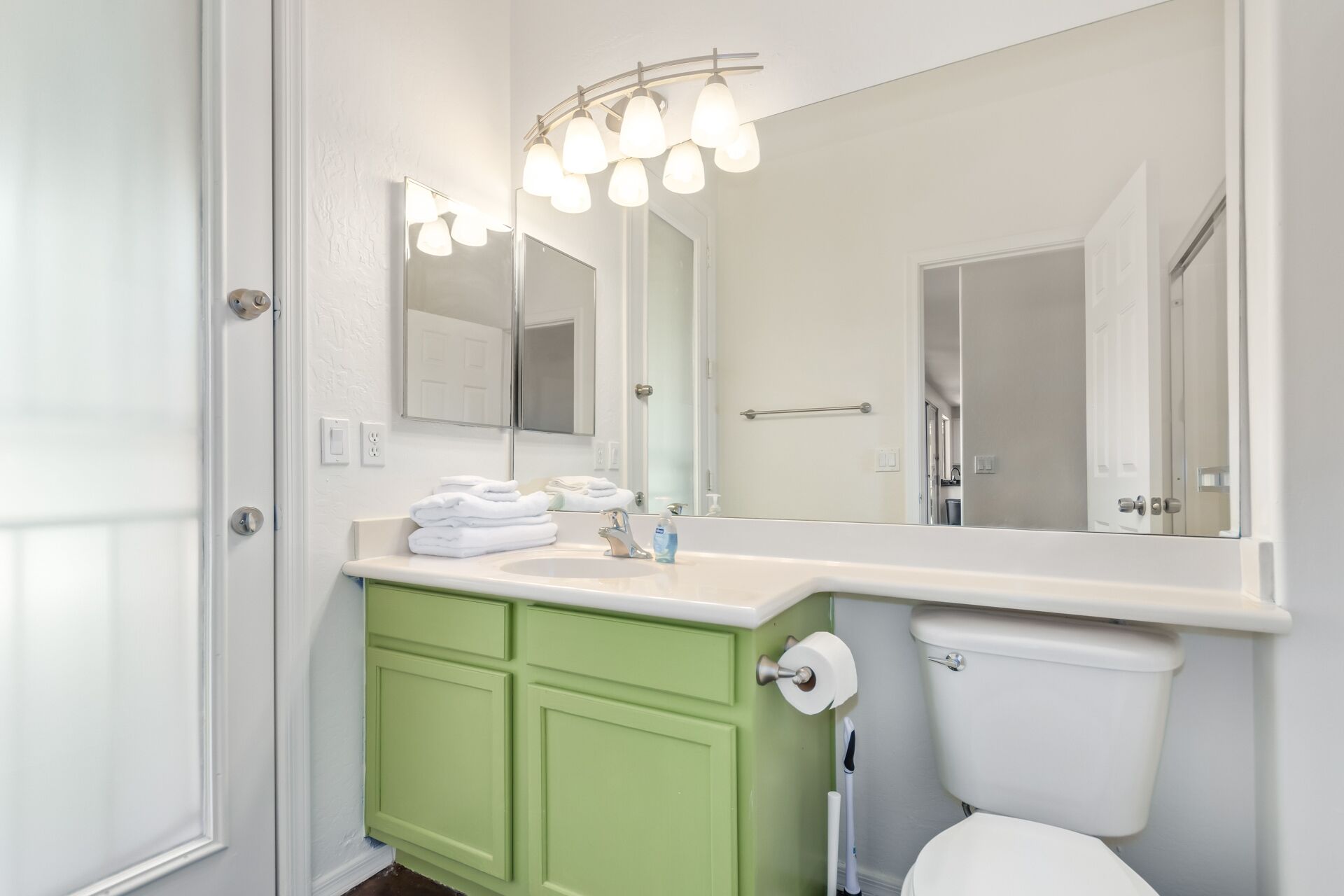 A bathroom with a white sink, green cabinets, a toilet, and a mirror with lights.