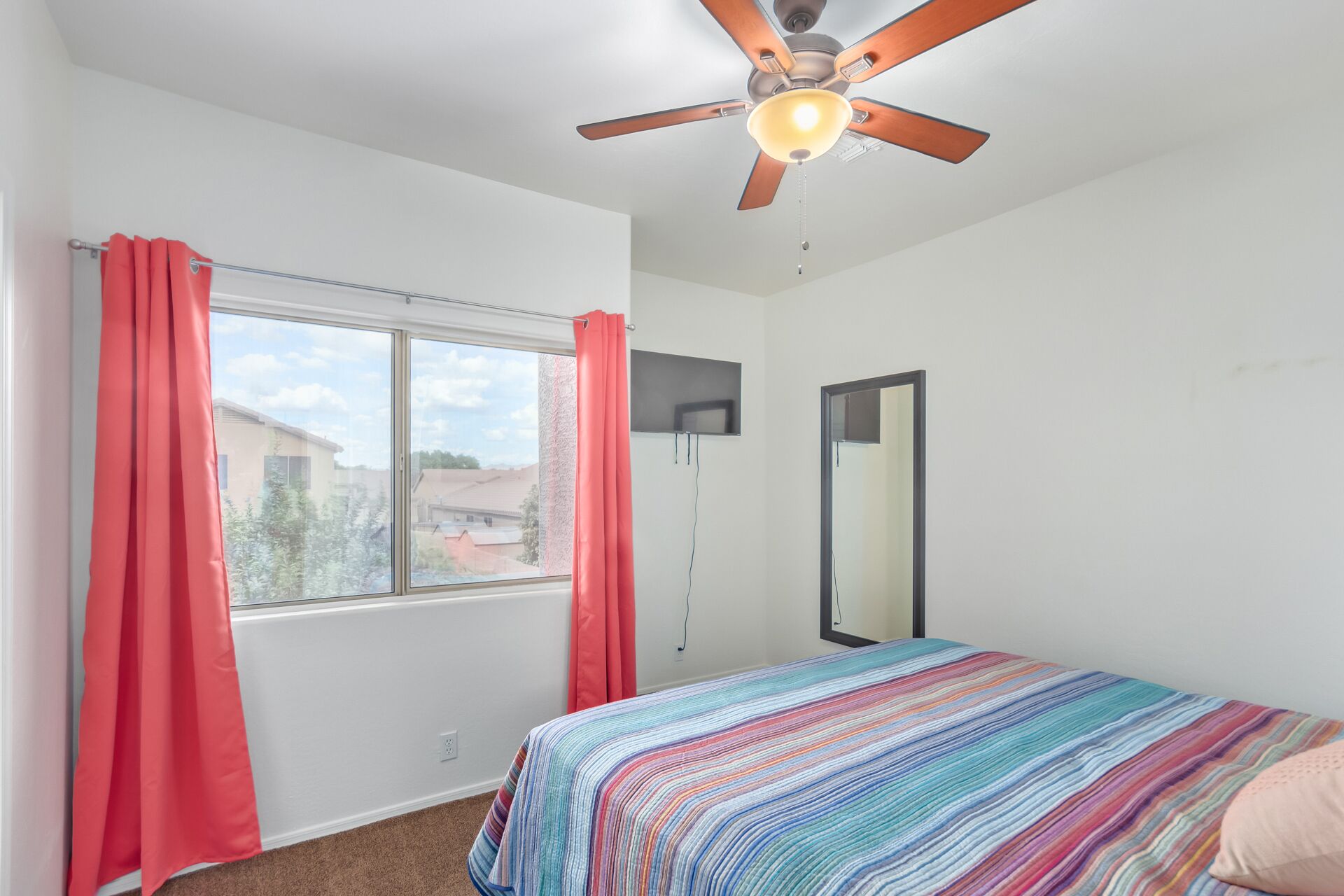 A bedroom with a bed, a ceiling fan, a window with curtains, and a mirror.