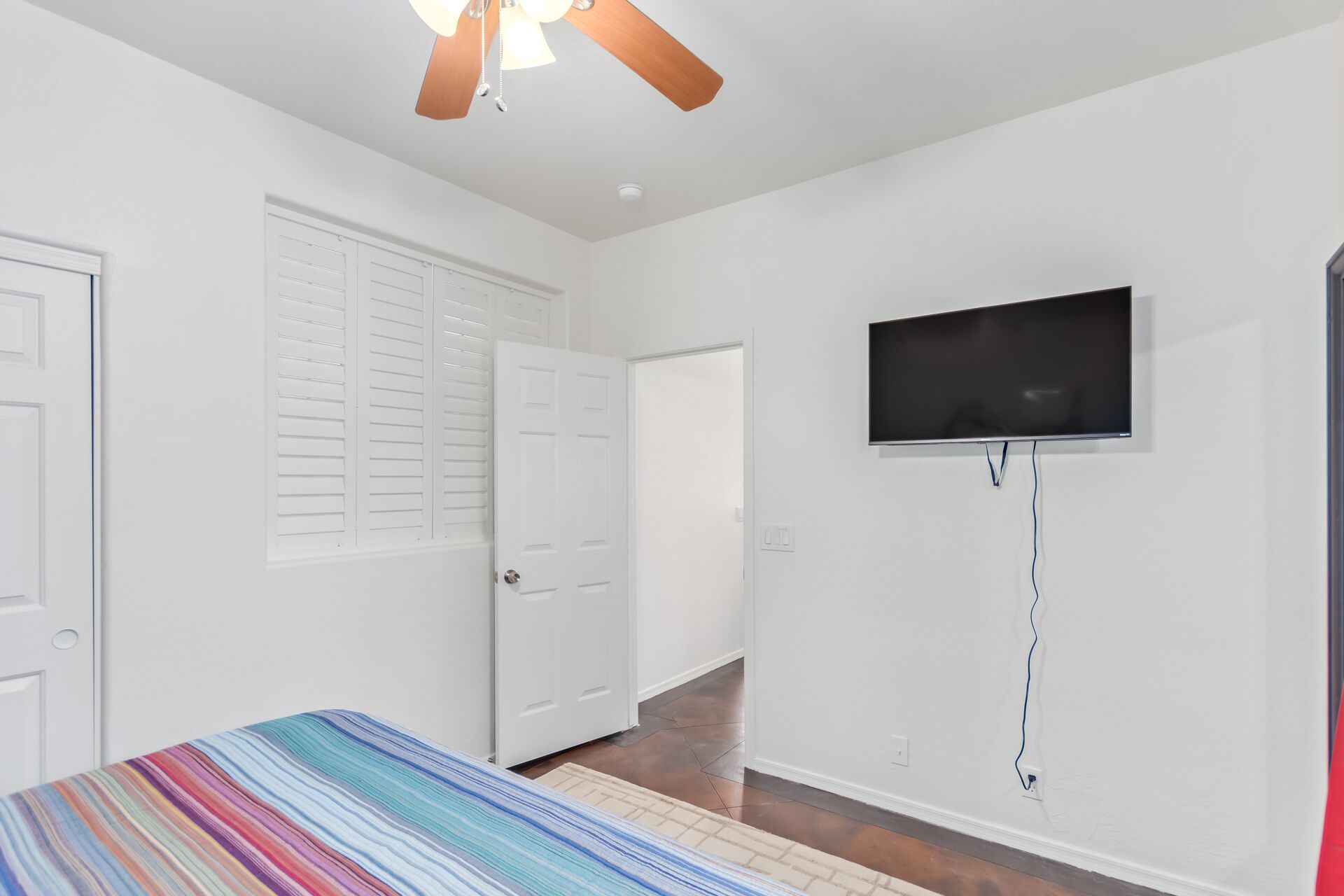 Ein Schlafzimmer mit Bett, Deckenventilator, an der Wand montiertem Fernseher und einer Tür zu einem anderen Raum.