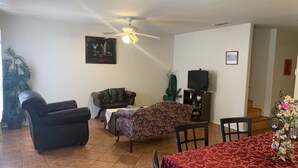 A living room with a ceiling fan, a sofa, a floral armchair, a TV stand, and a dining table with a red tablecloth.