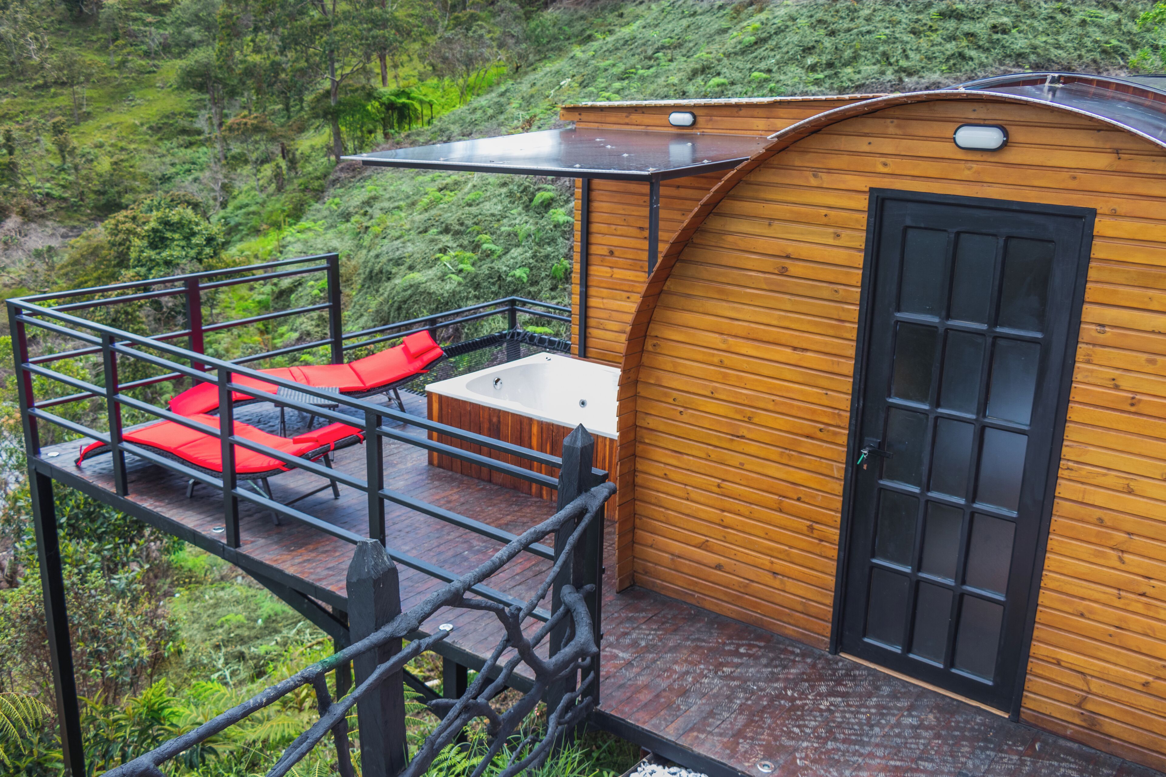 Una cabaña de madera con puerta negra, una pequeña terraza con asientos rojos y vistas a una ladera verde exuberante.