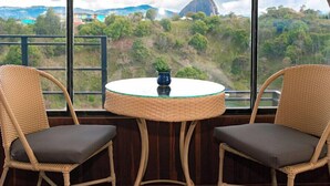 A room with a large arched window offering a view of a mountainous landscape. There is a round table with a blue vase in the center, flanked by two wicker chairs with grey cushions. - Glamping The Mountain (Guatapé)