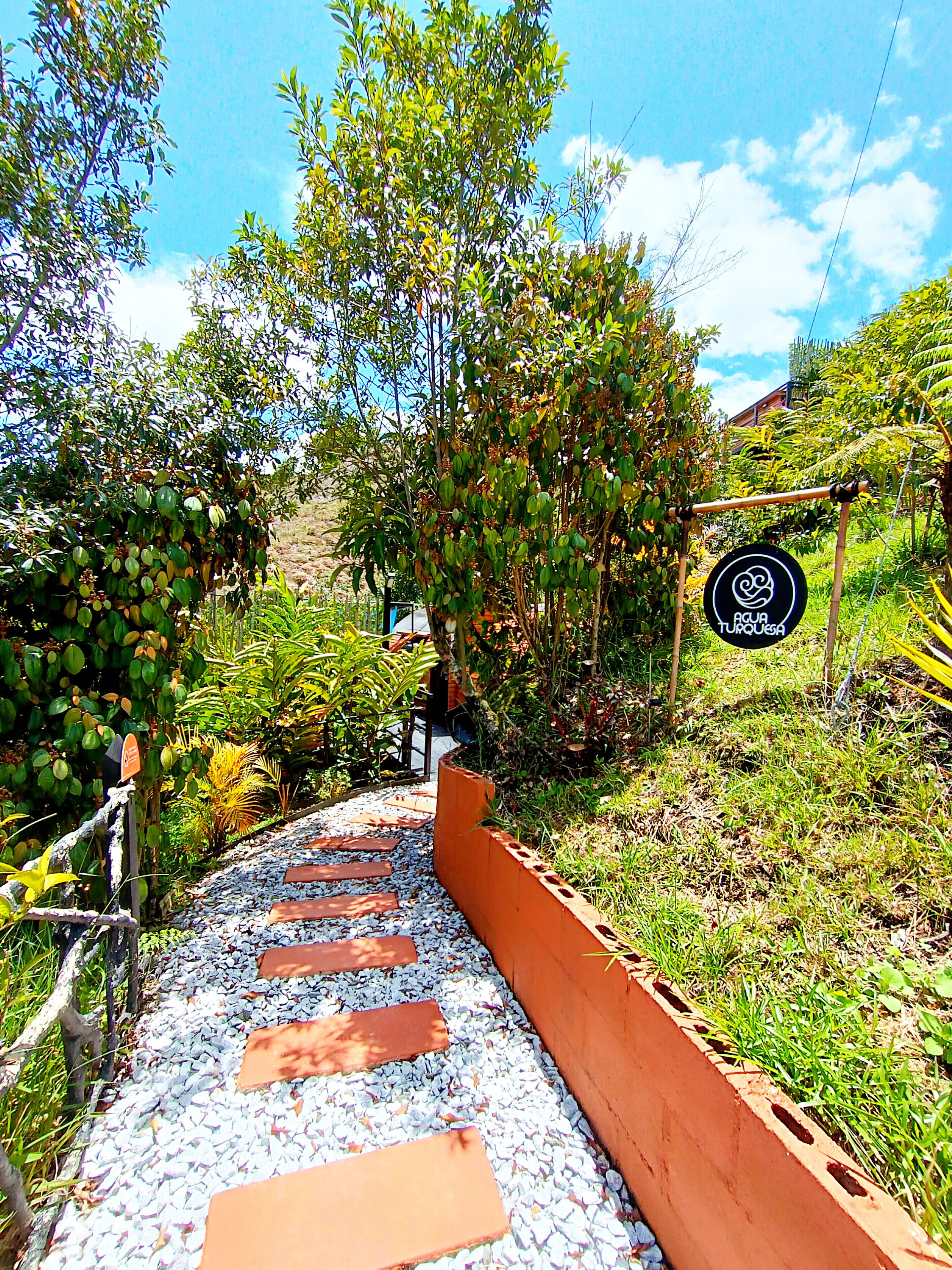 Uma passagem com pedras no caminho serpenteia por um jardim com diversas plantas e uma placa com a inscrição 'AQUA TURQUESA'.