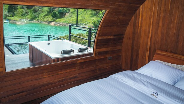 A wooden cabin with a large window overlooking a body of water and a deck area with a hot tub. - Glamping The Mountain (Guatapé)