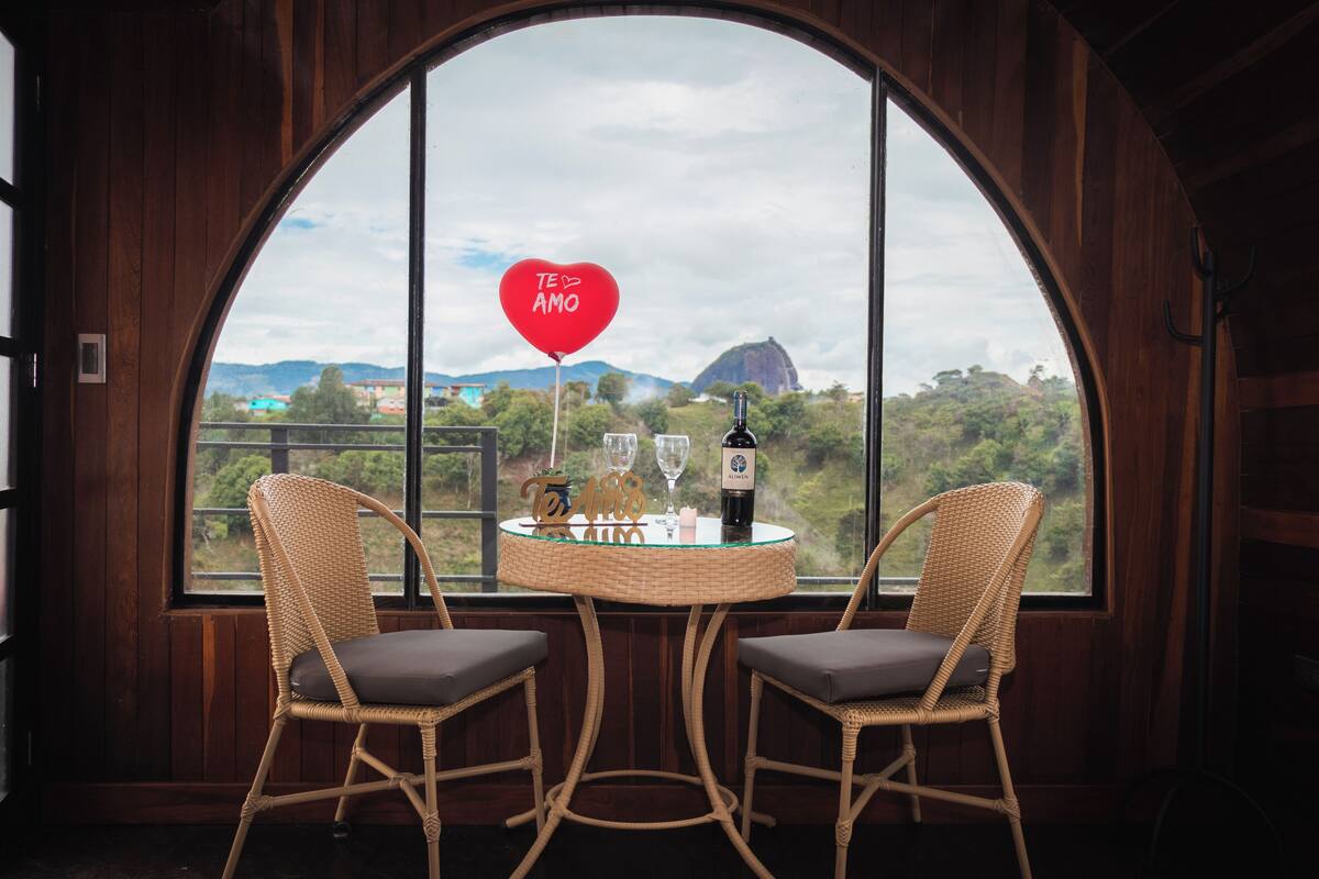 Une pièce avec deux fauteuils en osier et une petite table, une grande fenêtre en arc de cercle et une vue sur une montagne.