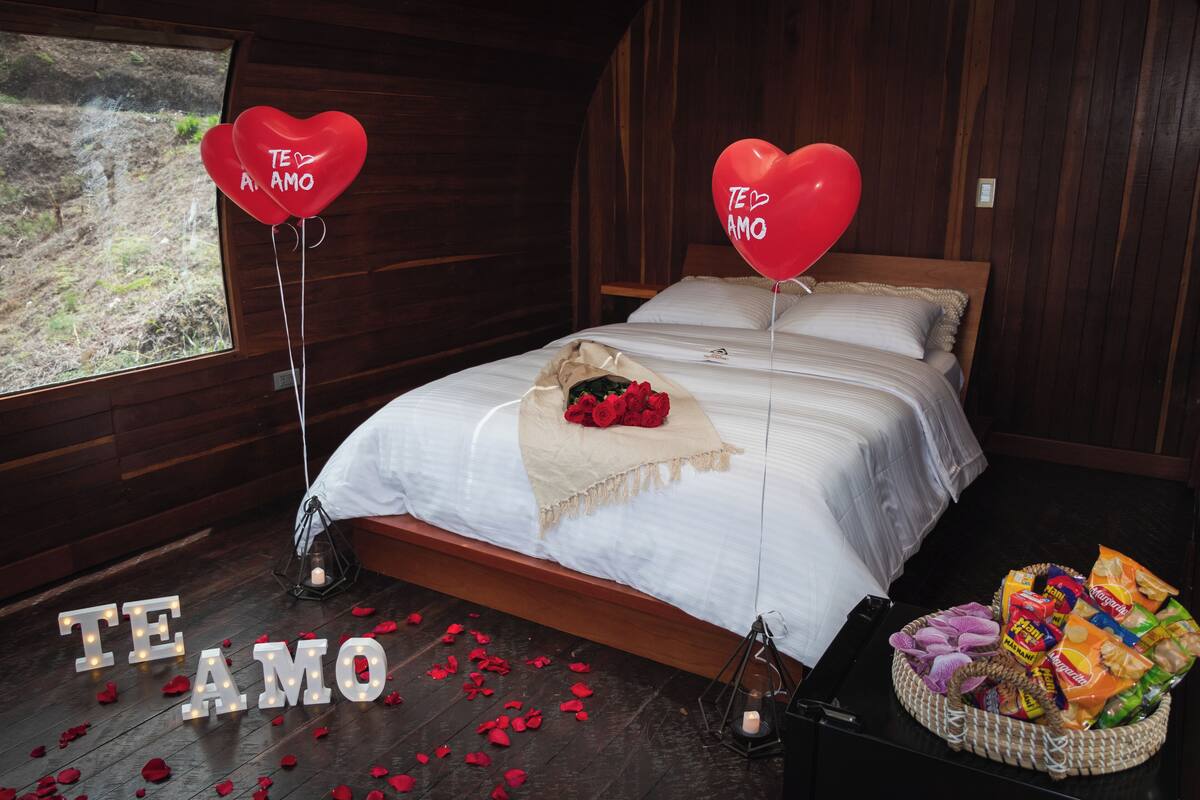 Une chambre avec un lit en bois, du linge de lit blanc, un bouquet de roses rouges, des ballons en forme de cœur et un panier rempli de collations.