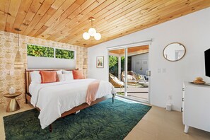 A bedroom with a wooden ceiling, a bed with white bedding and orange pillows, a green rug, and a sliding glass door leading to an outdoor area.