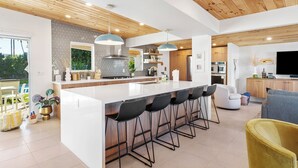 A modern kitchen with a large island, bar stools, and a dining area with a sofa and chairs. - NEW! Fully remodeled MCM w/mtn views, pool, & large yard! Designed by HGTV star! (Palm Springs)