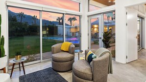 A modern living room with a large sliding glass door leading to a pool area, a cozy seating area with two armchairs, and a clock on the wall. - NEW! Fully remodeled MCM w/mtn views, pool, & large yard! Designed by HGTV star! (Palm Springs)